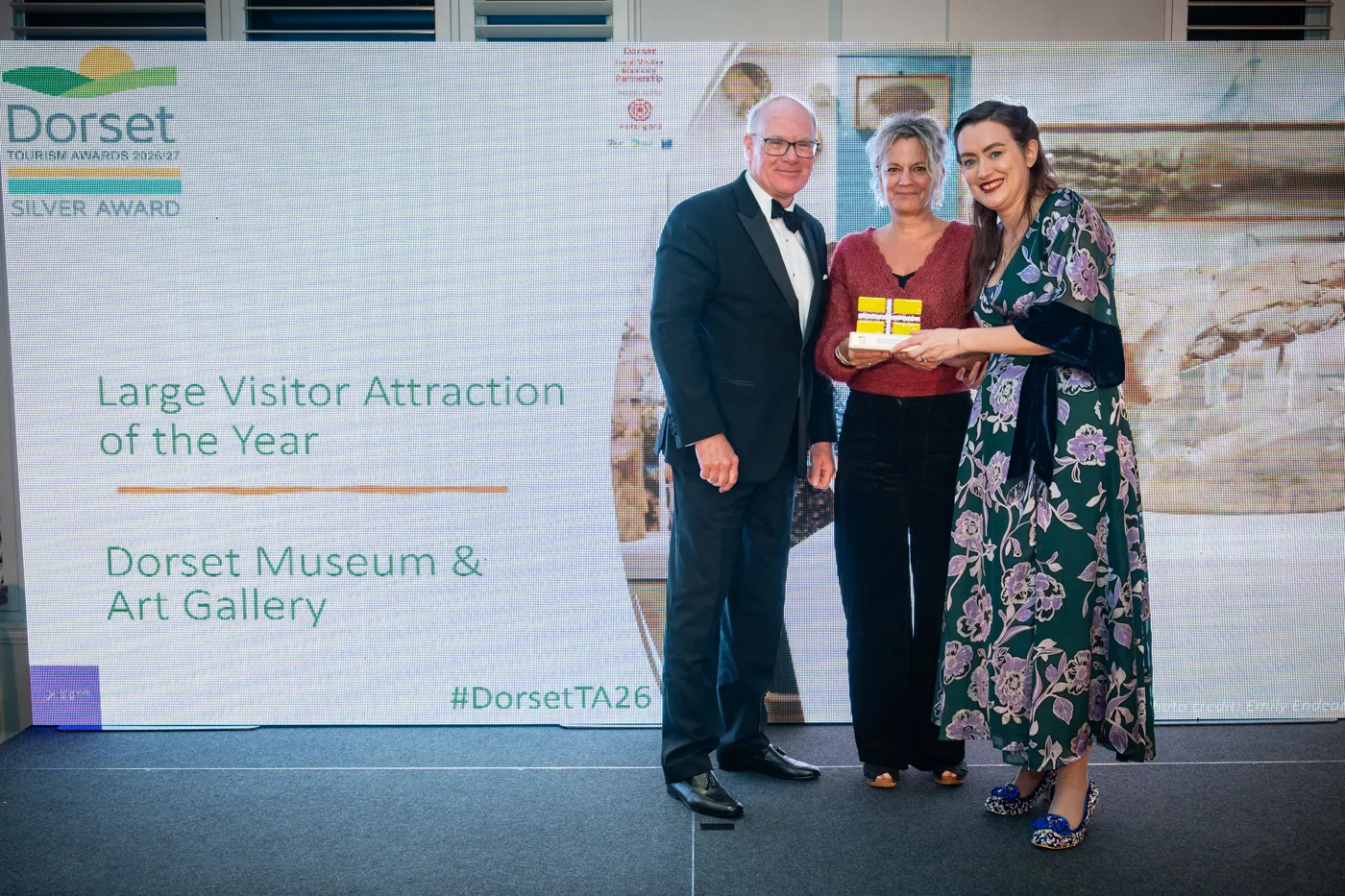 Three people on stage holding a yellow award block in front of a screen reading Large Visitor Attraction of the Year, Dorset Museum and Art Gallery, Silver Award