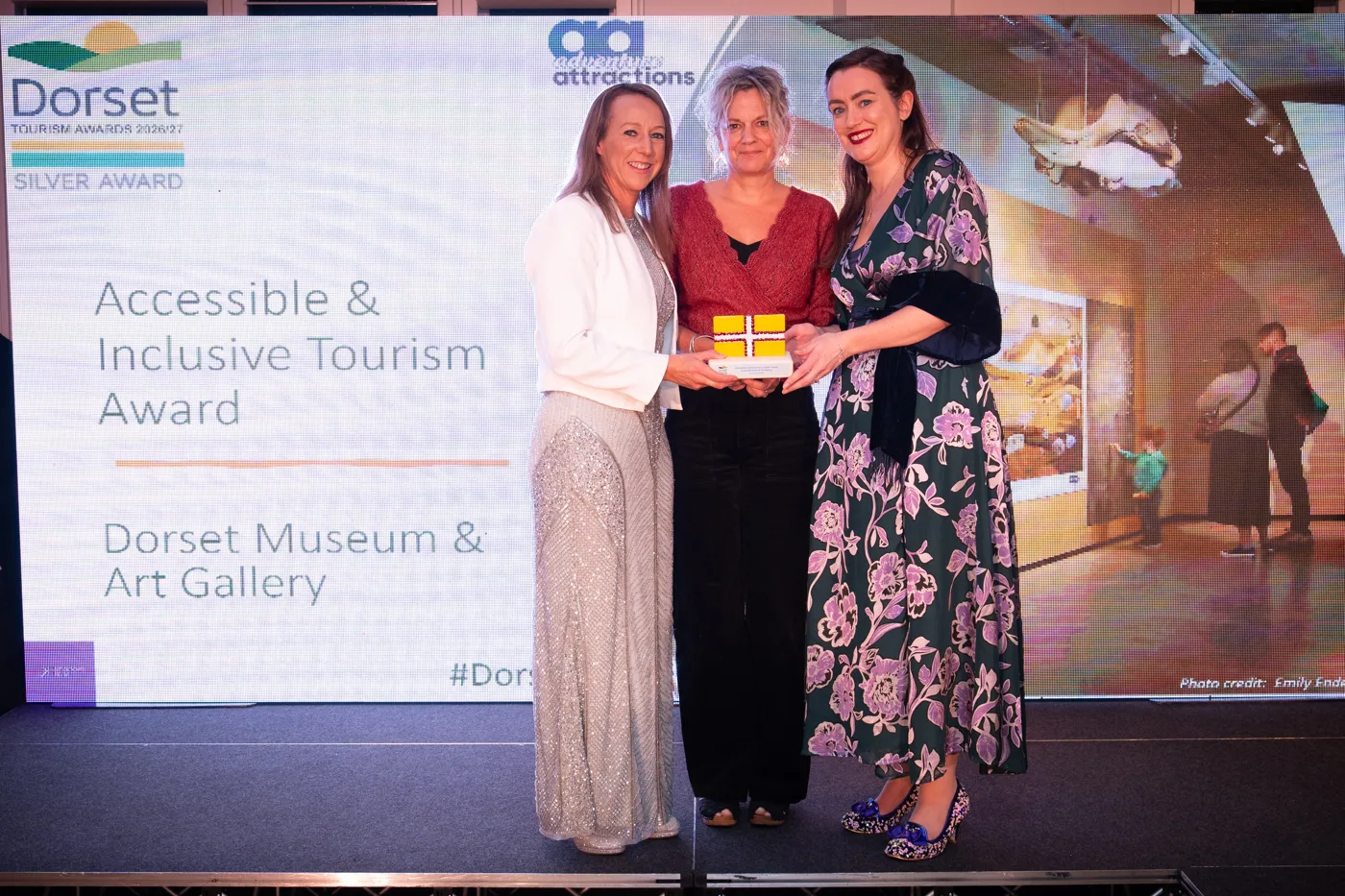 Three people on stage holding a yellow award block in front of a screen reading Accessible and Inclusive Tourism Award, Dorset Museum and Art Gallery, Silver Award