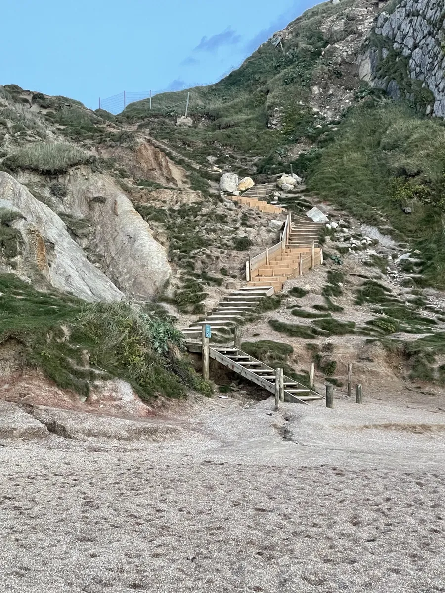 Timber steps zigzagging up a steep, grassy, eroded slope from the shingle beach at Durdle Door