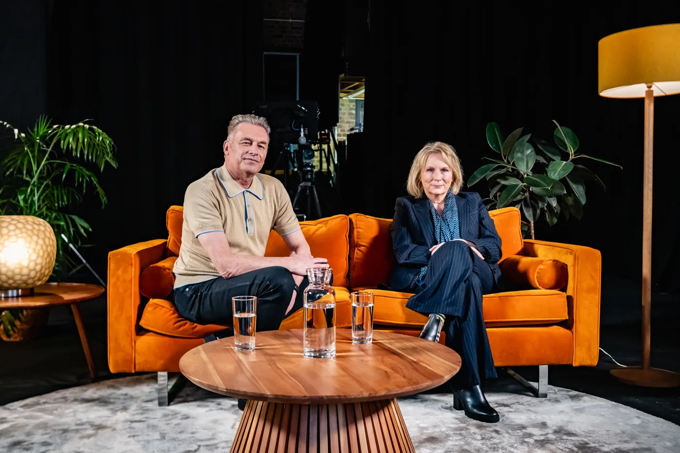 Two people seated on an orange sofa beside a round wooden table with water glasses, with a camera on a tripod behind them
