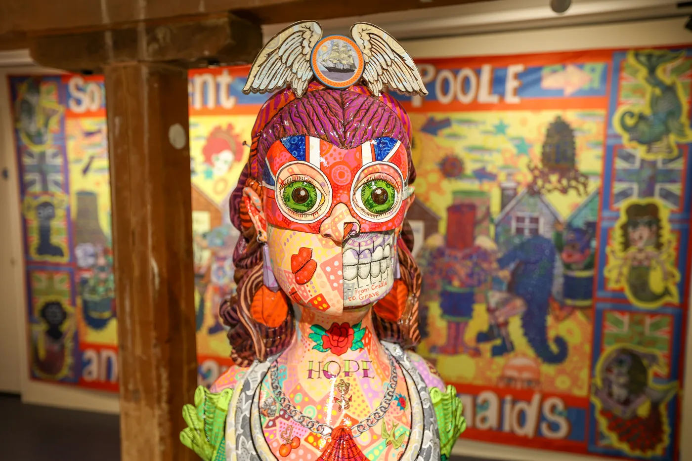 Close view of the painted masthead sculpture: a woman's face in a red mask with large green eyes, winged headdress with a ship, the word HOPE on the chest, in front of a large colourful Poole mural