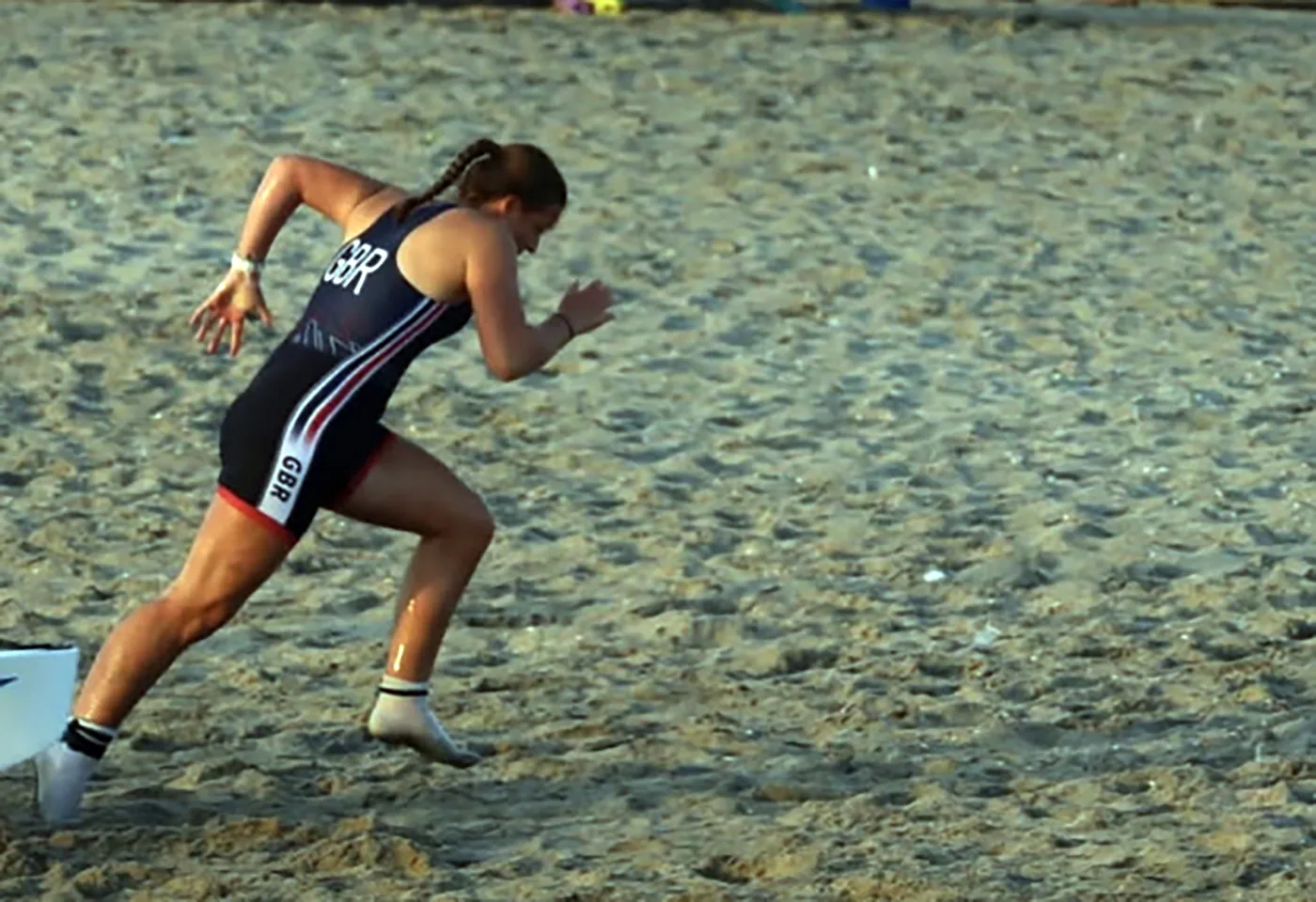 Lexi Gill in a navy GBR racing suit sprinting across sand away from a boat