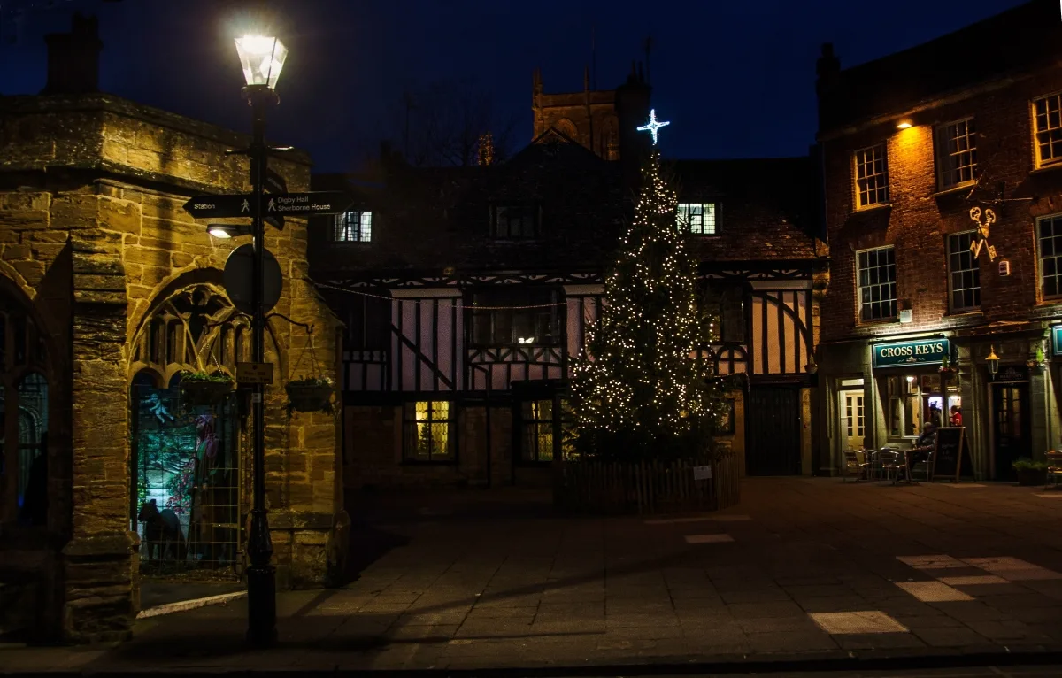 A Christmas tree strung with white lights at the foot of Cheap Street in Sherborne at night, beside the Cross Keys pub