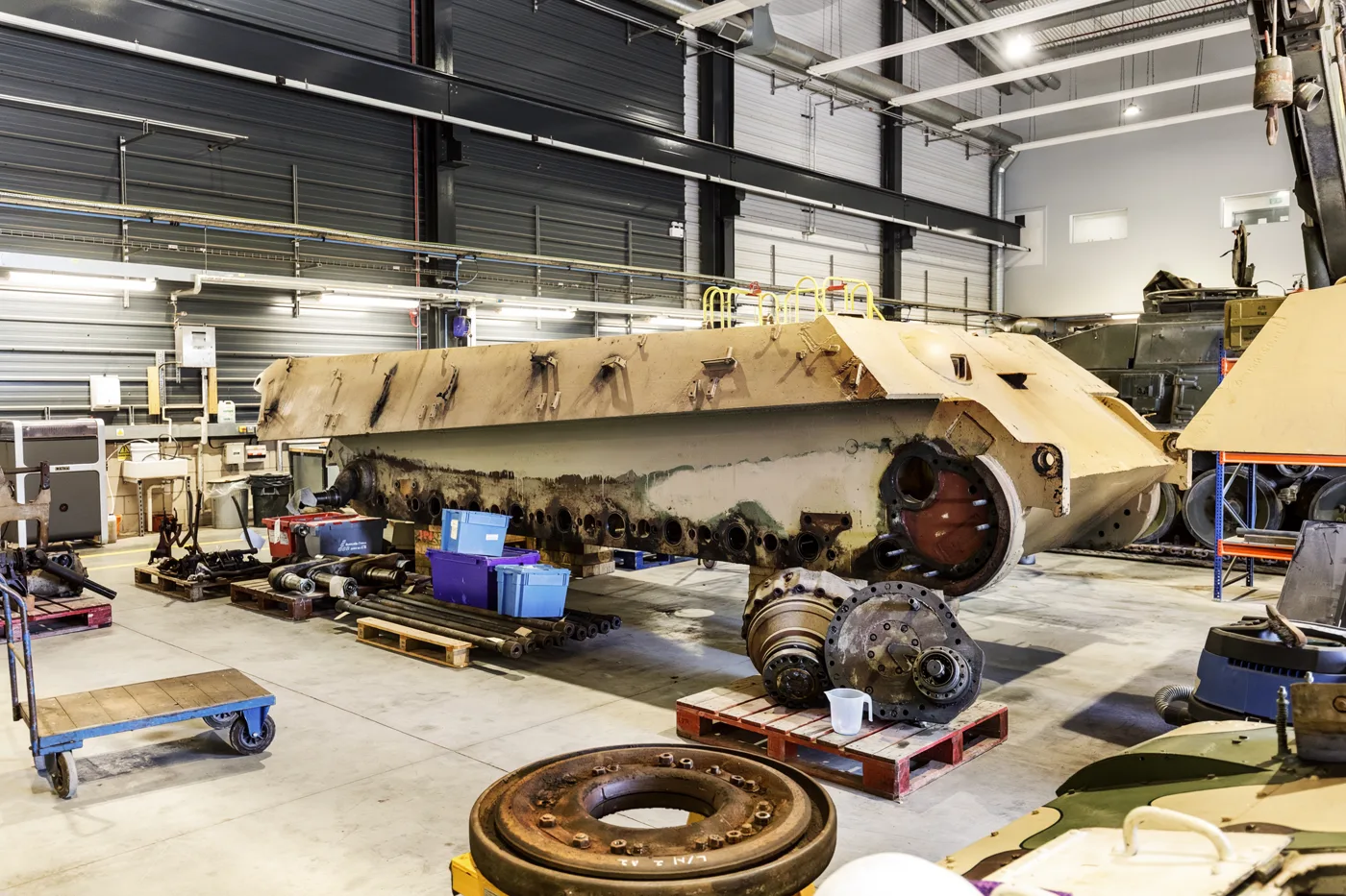 The stripped hull of King Tiger V2 in the Tank Museum workshop at Bovington, with parts laid out on pallets beside it.