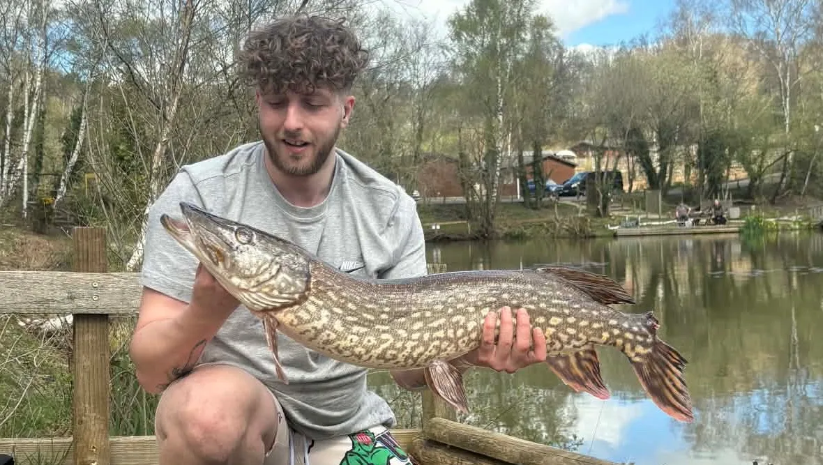 Martin James holding a pike beside a fishing lake