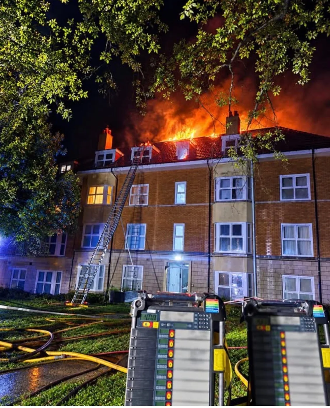 Flames breaking through the roof of Keel House at night, with a ladder against the building and hose lines across the grass