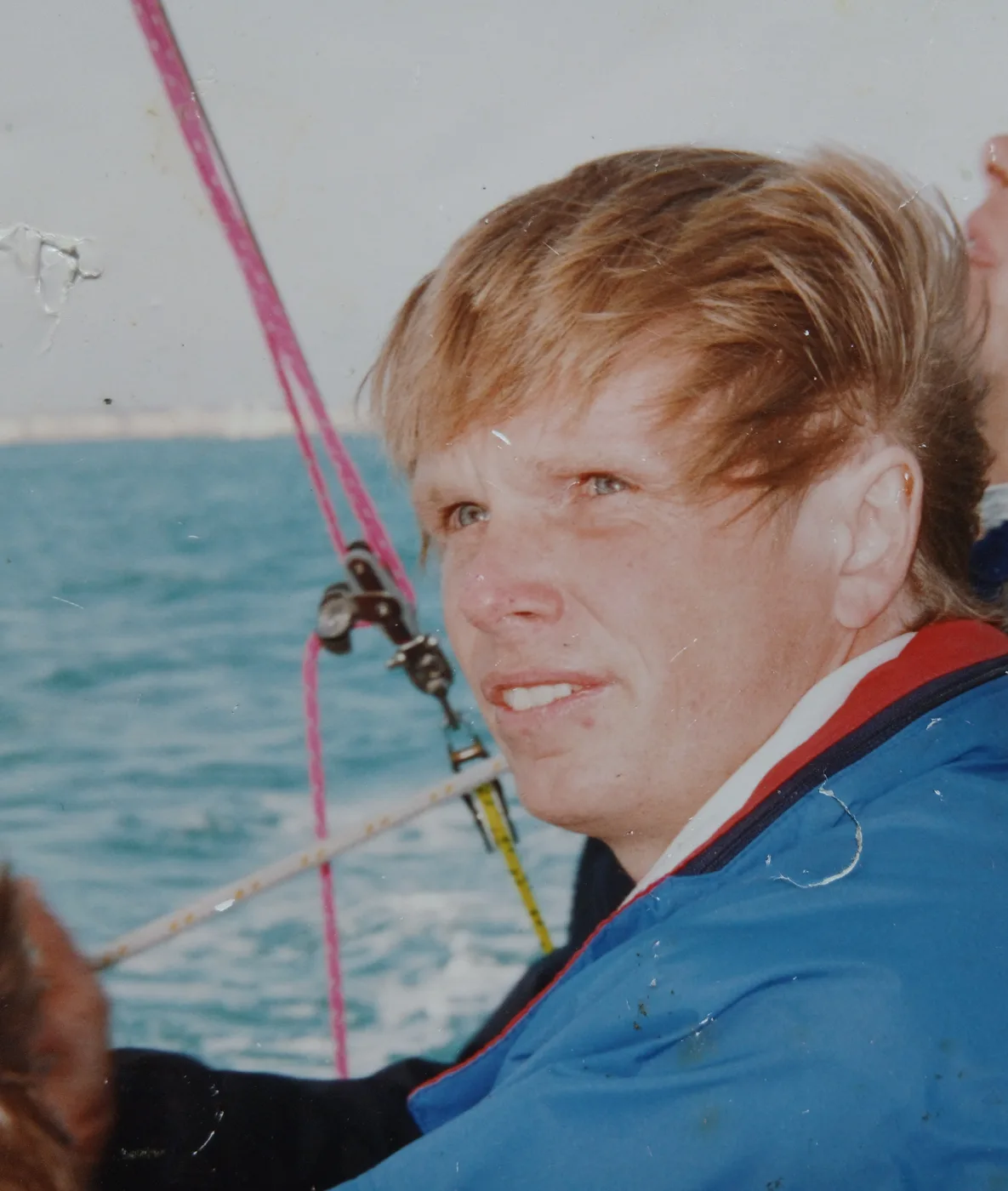 Terry Ridyard in a blue and red jacket aboard a boat, with rigging lines and the sea behind him